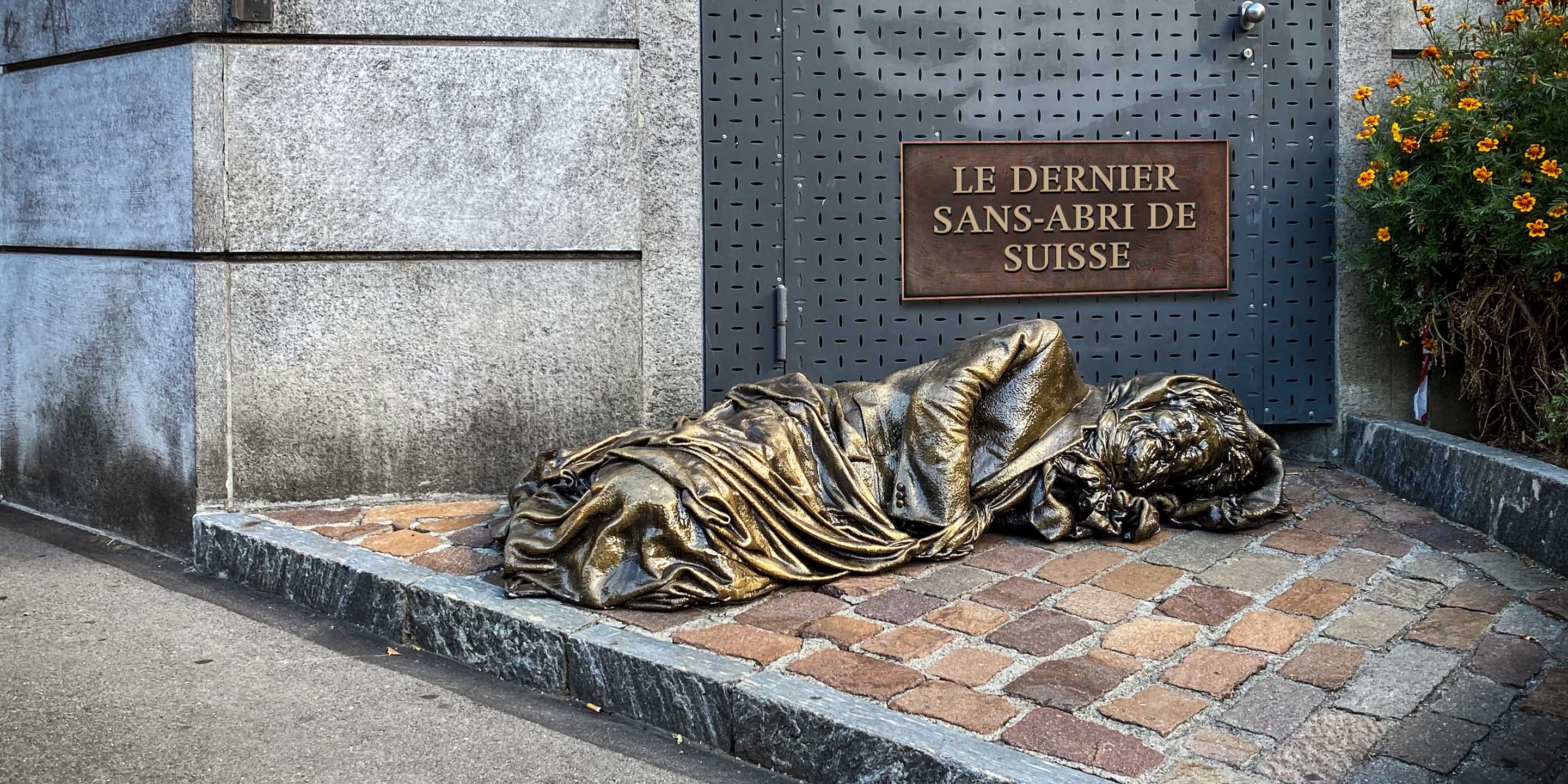 Eine Heilsarmee-Offizierin sitzt neben einer Statue eines Obdachlosen. Oberhalb der Statue steht an der Wand der Schriftzug 