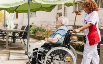 Frau Fuss wird im Rollstuhl von einer Pflegerin durch den Aussenbereich geschoben